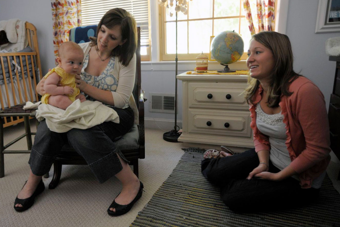 A woman is gently holding a baby in a warm, comfortable room while chatting with April.