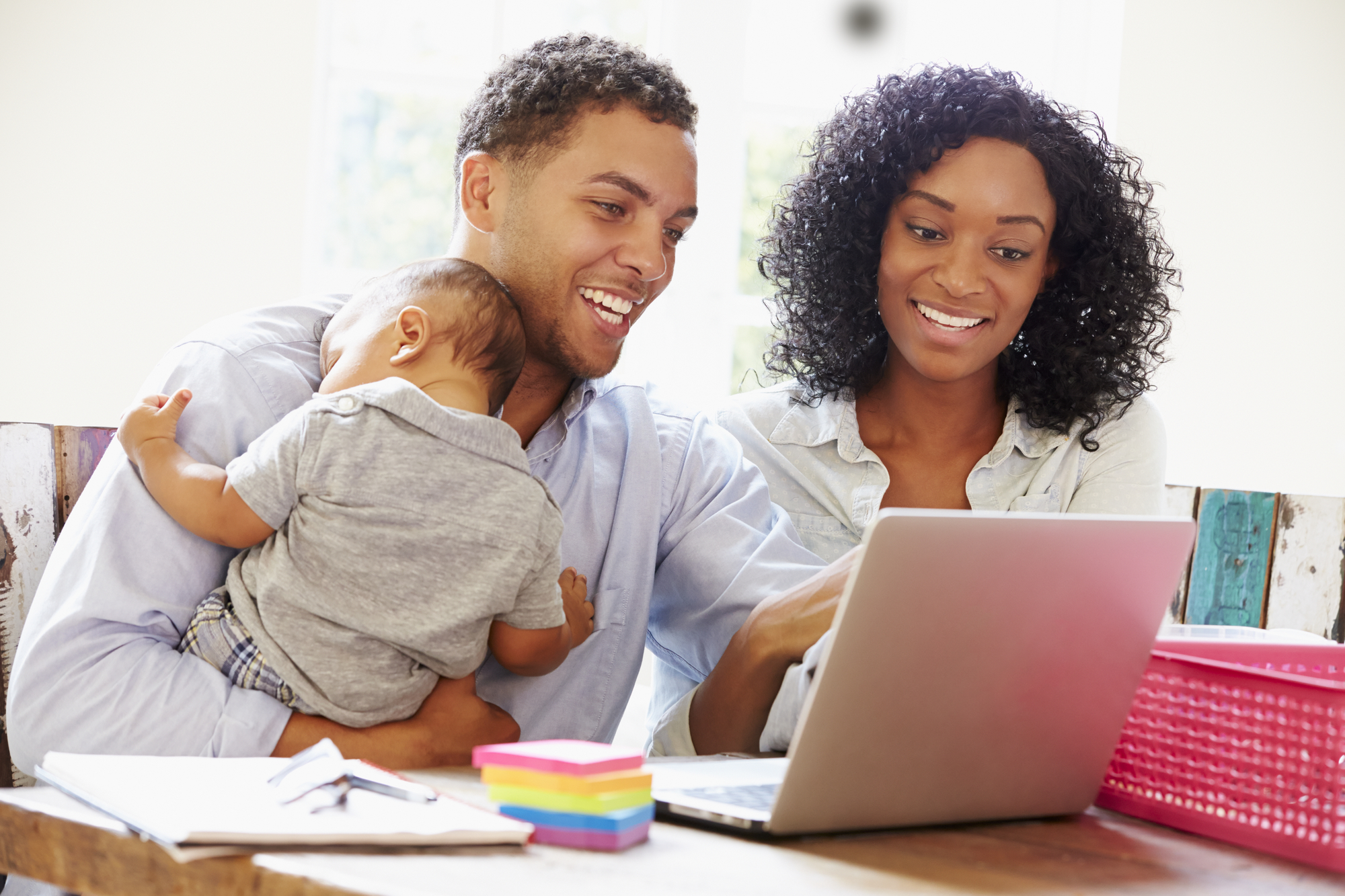 Happy young parents with baby using laptop at dining room table.